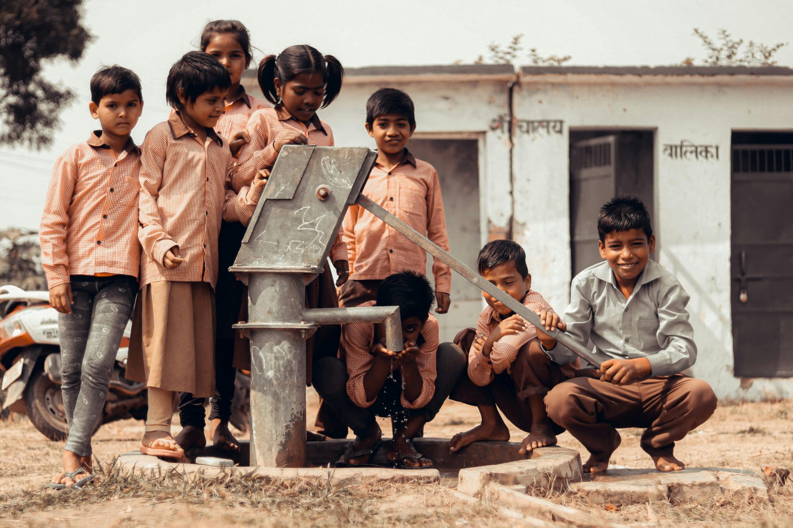 Home A group of Indian children gather joyfully around a manual water pump outdoors.
