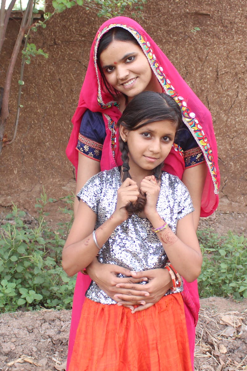 Home A smiling woman and girl in colorful traditional Indian attire, embracing warmly outdoors.